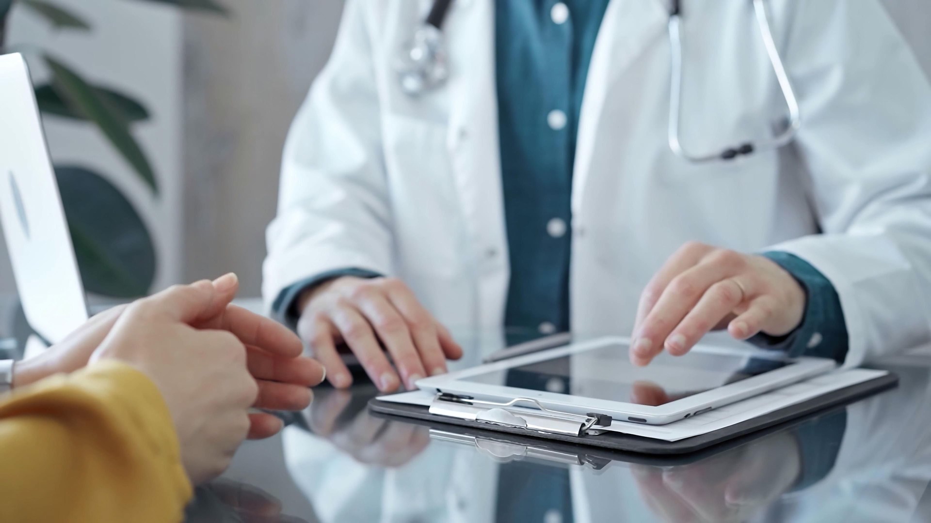 Doctor and patient consultation in clinic. Patient woman is gesturing over a glass desk while a physician is using tablet computer making some notes, close up of hands. Medicine and health care